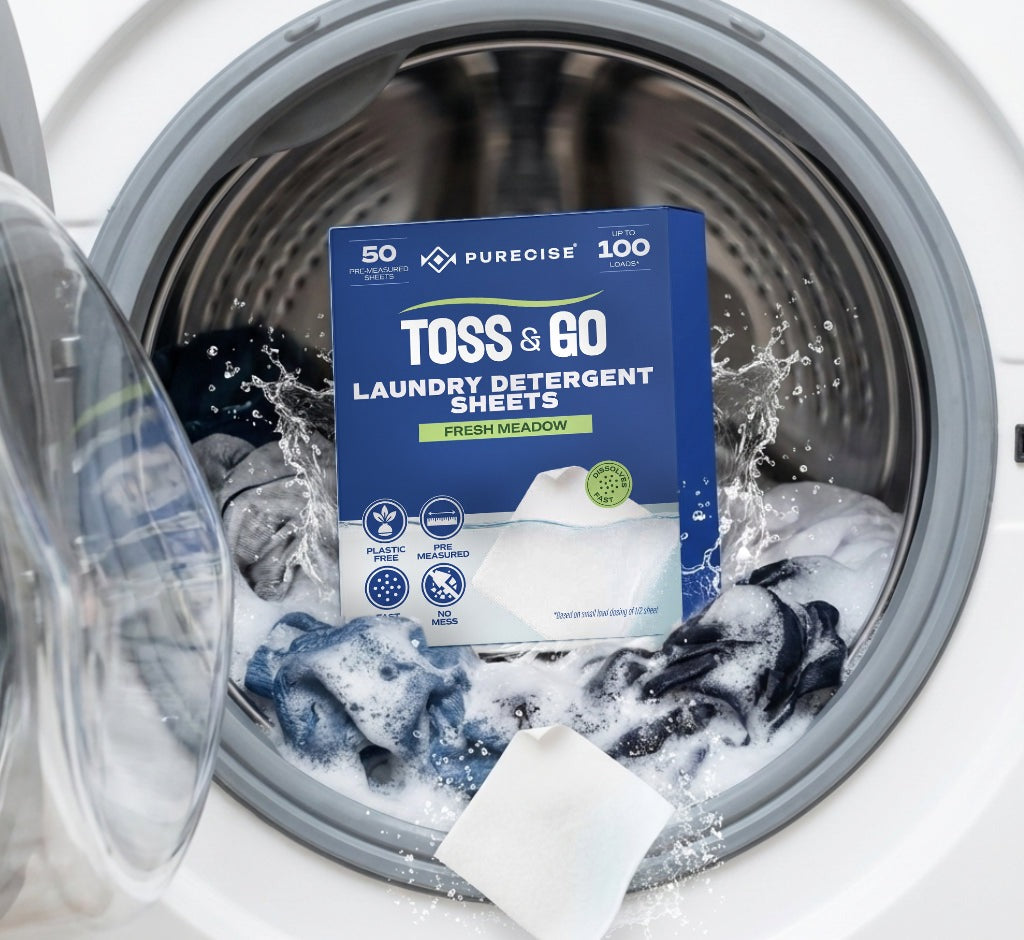 A Purecise Toss & Go box of 50 laundry detergent sheets, Fresh Meadow, in a front-load washing machine drum with sudsy laundry